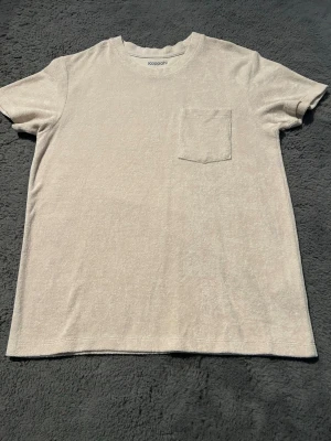 Beige t-shirt med bröstficka Kappahl - En stilren beige t-shirt från Kappahl med rund halsringning och en enkel bröstficka. T-shirten har korta ärmar och är tillverkad i ett mjukt material som känns skönt mot huden. Perfekt för en avslappnad look.