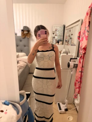 Vit och svart långklänning med blommönster - Långklänning i vit och svart med blommönster och stickad struktur. Klänningen har smala axelband och en kroppsnära passform som framhäver siluetten. Perfekt för sommarens varma dagar och snygg att styla med sandaler eller sneakers.