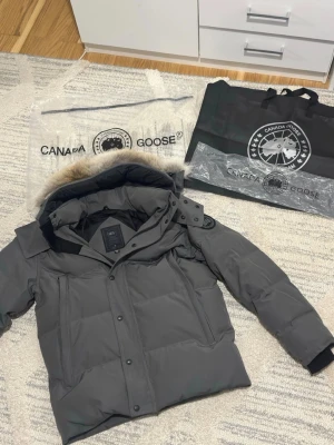 Grå Canada Goose Wyndham - Säljer en stilren grå Canada Goose vinterjacka i mycket fint skick. Har varm dunfyllning, huva med pälskant och klassisk Canada Goose-logga på ärmen. Perfekt för kalla dagar och för dig som vill ha en både snygg och riktigt varm jacka. Kvitto följer med