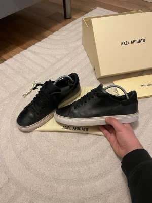 Svarta Axel Arigato sneakers - Stilrena svarta sneakers från Axel Arigato i läder med klassisk låg profil och vit sula. Diskret logga på sidan och svarta skosnören. Kommer med originalkartong och dustbag. Perfekta för dig som gillar minimalistisk och clean stil.