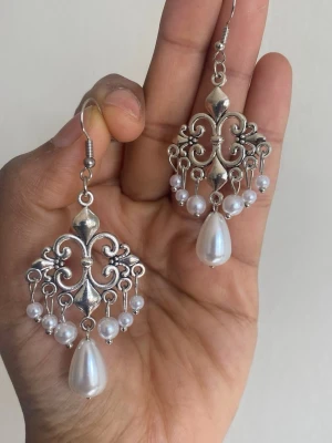 Silvrig färgat örhängen med pärlor - Unika silvriga örhängen med vackra detaljer och hängande vita pärlor. Örhängena har en elegant, ornamenterad design och stora droppformade pärlor längst ner. Perfekta för att ge din stil en extra touch av glamour.