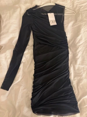 Zara svart mesh one shoulder klänning - Aldrig använd kort svart klänning i mesh med one shoulder-design och lång ärm. Klänning har ickså meshdeyalj vid magen. Köpt för 399kr, prislappen sitter kvar