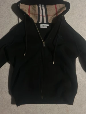 Svart Burberry hoodie med dragkedja - Svart hoodie från Burberry med dragkedja och klassiskt rutigt foder i huvan. Guldiga detaljer på dragkedja och snören. Mjuk bomullskänsla och snygg passform, perfekt för en stilren och lyxig look. M i England storlek den passar på xs mycket bättre 
