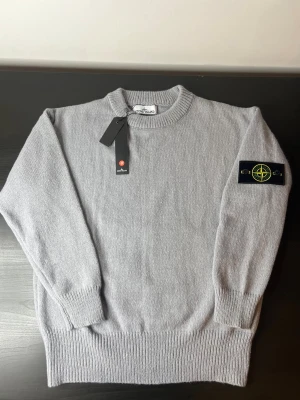 Grå stickad tröja från Stone Island - Snygg grå stickad tröja från Stone Island med klassisk rund hals och ribbade muddar. Tröjan har den ikoniska Stone Island-loggan på vänster ärm och är tillverkad i mjukt material som passar perfekt till höst och vinter.