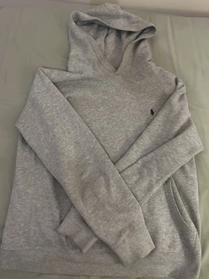 Grå hoodie från Polo Ralph Lauren - Snygg gråmelerad hoodie från Polo Ralph Lauren med klassisk huva och liten broderad logga på bröstet. Mjuk och skön bomullsblandning, perfekt för en avslappnad stil. Har känguruficka framtill och ribbade muddar vid ärmslut och nederkant.