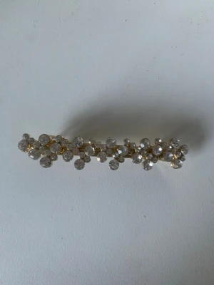 Glittrig hårklämma med stenar - Snygg hårklämma i guldmetall dekorerad med klara glittrande stenar i blommönster. Perfekt accessoar för att ge frisyren extra bling och stil. Klämman har en avlång form och passar till många olika looks.