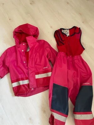 Rosa regnjacka+Regnbyxor  från Didriksons - Snygg rosa regnjacka från Didriksons samt regnbyxor  med avtagbar huva och reflexdetaljer nedtill. Jackan har fleecefoder på insidan för extra värme och tryckknappar framtill. Perfekt för regniga dagar och enkel att matcha med andra plagg.   Allt fleecefodrat Fleecefodrad