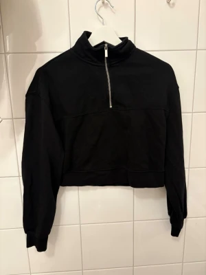 Svart cropped hoodie från H&M - Säljer en svart cropped hoodie från H&M med halv dragkedja framtill och hög krage. Hoodien har långa ärmar med muddar och är tillverkad i mjukt sweatshirtmaterial. Perfekt för en avslappnad och trendig look.
