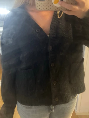 Svart stickad kofta med knappar - Mysig svart stickad kofta med v-ringning och stora knappar framtill. Koftan har ribbade muddar vid ärmslut och nederkant samt två fickor framtill. Perfekt att slänga över en t-shirt eller linne för en avslappnad look.