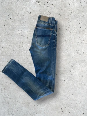 Blå slim fit jeans från Nudie Jeans - Säljer ett par blå slim fit jeans från Nudie Jeans med klassisk femficksdesign och snygga fades. Jeansen har en mörkblå tvätt med ljusare partier och kontrastsömmar. Perfekta för en avslappnad och trendig look. Pris kan diskuteras 