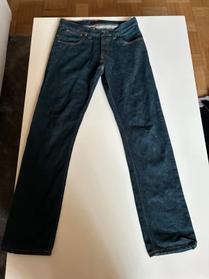 Mörkblå jeans från Nudie Jeans - Säljer ett par mörkblå raka jeans från Nudie Jeans med klassiska orange sömmar och broderade bakfickor. Jeansen har fem fickor och är tillverkade i denim. Perfekta för dig som gillar en tidlös och stilren look.