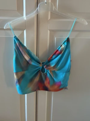 Färgglad linnetopp med ringdetalj från Bershka - Snygg och färgglad linnetopp med smala axelband och en cool ringdetalj framtill från Bershka. Toppen har ett mönster i blått, rosa och orange toner och är perfekt för varma dagar. Materialet känns lätt och luftigt. I fint skick. Storlek M. Croppad topp. 