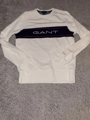 Vit sweatshirt från GANT - Snygg vit sweatshirt från GANT med marinblått band över bröstet och logga i vitt. Tröjan har rund halsringning, långa ärmar och ribbade muddar. Perfekt för en avslappnad och stilren look. S ny pris 1000kr+, MITT PRIS 450 