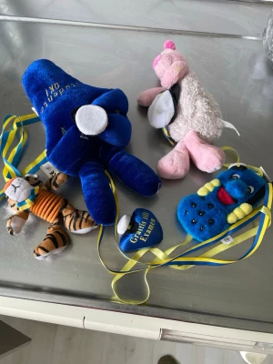 Studentnallar och mjukisdjur med band - Fyra olika mjukisdjur med blågula band, perfekta till studentfirande. En blå elefant med text, en rosa flamingo, en tiger med orange tröja och en blå mask med gula prickar. Materialet är mjuk plysch och färgerna är blå, rosa, orange och gul.
