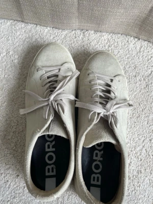 Vita sneakers från Björn Borg - Stilrena vita sneakers från Björn Borg i mocka med klassisk låg profil och vita skosnören. Skorna har en enkel design med rund tå och vit sula, perfekta för en clean och avslappnad look.
