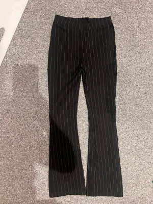 Svarta kostymbyxor med vita ränder - Snygga svarta kostymbyxor med smala vita kritstrecksränder och utsvängda ben. Byxorna har hög midja och klassisk passform som ger en stilren look. Perfekta för dig som gillar en trendig och tidlös stil.