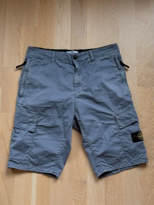 Grå cargoshorts från Stone Island - Säljer ett par grå/blå cargoshorts från Stone Island med klassisk patch på benet. Shortsen har flera fickor, bälteshällor och knappstängning. Perfekta för sommaren och har en avslappnad passform. Funkar perfekt för båda män och kvinnor! Originellt pris är 3399kr. Säljer dessa för 850kr. 