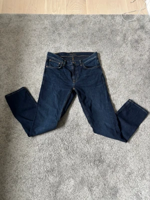 Mörkblå jeans från Nudie Jeans - Säljer ett par klassiska mörkblå jeans från Nudie Jeans med raka ben som aldrig är använda. Jeansen har kontrastsömmar och en patch med logga bak i midjan. Tillverkade i slitstark denim, perfekta för en clean och tidlös look. Priset går att diskutera. Vid intresse är det bara att höra av sig
