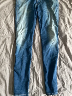 Replay jeans anbass - Snygga blå slim fit jeans med ljusa slitningar framtill och klassiska fem fickor. Jeansen har en enkel bakficka med diskret sömnad och är tillverkade i ett mjukt denimtyg som ger en bekväm känsla. Perfekta för en avslappnad och trendig stil.