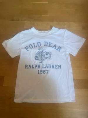 Vit Polo Bear t-shirt Ralph Lauren - Vit t-shirt från Ralph Lauren med Polo Bear-tryck och texten 'Ralph Lauren 1967' i blått framtill. Klassisk rund hals och korta ärmar. Perfekt för en avslappnad och cool stil.