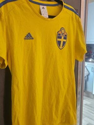 Svensk landslagströja Adidas gul - Gul svensk landslagströja från Adidas med blå detaljer och SVFF-emblem på bröstet. Tröjan har korta ärmar, rund hals och klassiska Adidas-ränder på axlarna. Perfekt för fotbollsfans som vill stötta Sverige.