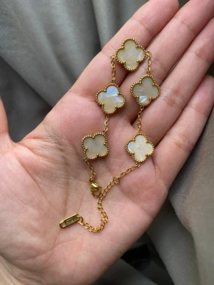 Guldpläterad armband  - Snyggt armband i guldton med fem vita fyrklöverformade hängen i pärlemor. Kedjan är tunn och har ett elegant lås. Perfekt accessoar för dig som gillar stilrena och trendiga smycken med en lyxig touch.