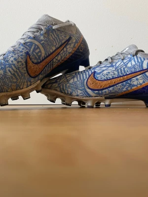 Nike Mercurial blå/guld fotbollsskor - Säljer ett par Nike Mercurial fotbollsskor i grått och blått med ett unikt mönster och guldigt Nike-märke på sidan. Skorna har snörning, låg profil och dobbar för spel på konstgräs. Ovandelen är i syntetmaterial för bra bollkontroll och passform. Pris kan diskuteras.
