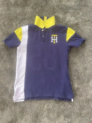 Blå och gul pikétröja från Tommy Hilfiger - Snygg pikétröja i blått, gult och vitt från Tommy Hilfiger. Tröjan har korta ärmar, klassisk krage i gult och blått samt TH-logga broderad på bröstet. Perfekt för en sportig och avslappnad look.