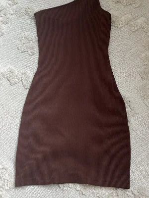 Brun oneshoulder kortklänning - Säljer en brun, ribbad oneshoulder-klänning i kort modell. Klänningen har en tight passform och är tillverkad i ett mjukt, stretchigt material som sitter snyggt på kroppen. Perfekt för dig som gillar stilrena och trendiga plagg.