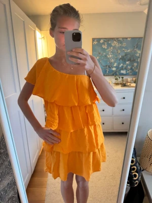 Orange volangklänning med asymmetrisk axel - Säljer en superfin orange kortklänning med flera volanglager och asymmetrisk axel. Klänningen har en luftig passform och är perfekt för varma dagar. Materialet känns lätt och svalt, och den har en trendig offshoulder-look. Nypris runt 1000 kr