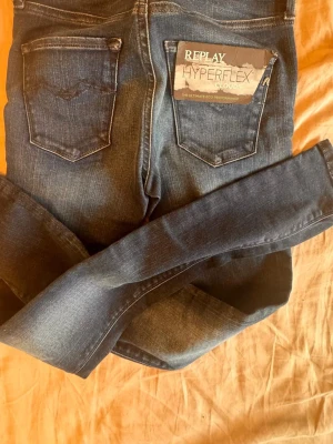 Replay Hyperflex blå jeans - Säljer ett par blå Replay Hyperflex jeans med klassisk femficksdesign och snygga slitningar. Jeansen är handgjorda och tillverkade i Italien, med stretchigt material som ger en skön passform. Perfekta för dig som vill ha både stil och komfort. Helt ny från Replay