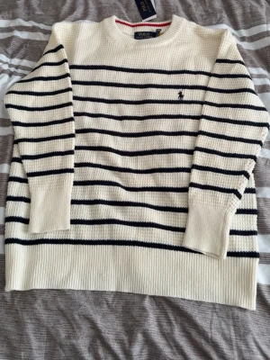 Randig stickad tröja från Polo Ralph Lauren - Klassisk stickad tröja i offwhite med svarta horisontella ränder från Polo Ralph Lauren. Tröjan har rund halsringning, långa ärmar och den ikoniska broderade loggan på bröstet. Tillverkad i mjukt material som ger en avslappnad men stilren look.