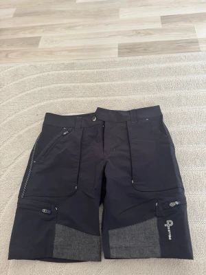 Svarta funktionsshorts från Pelle p - Säljer ett par svarta funktionsshorts från Pelle p med grå förstärkningar på bak och ben. Shortsen har dragkedjefickor fram och på benen, bälteshällor och en sportig look. Perfekta för utomhusaktiviteter och äventyr.