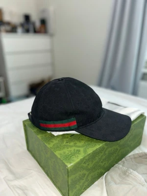Svart Gucci Keps!  dustbag, Kvitto och låda ingår. - Priset går att diskuteras! Snygg svart keps från Gucci med diskret GG-mönster i tyget och klassisk röd och grön rand på sidan. Kepsen har böjd skärm och justerbar rem baktill. Perfekt accessoar för att lyfta din streetwear-look.