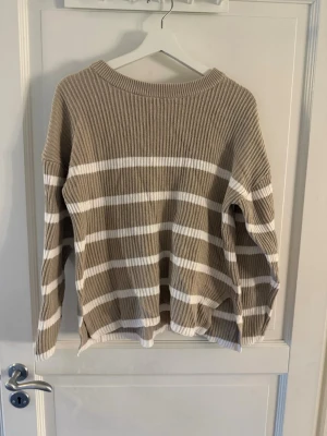 Beige och vit randig stickad tröja - Stickad tröja i beige med vita horisontella ränder. Tröjan har rund halsringning, lång ärm och ribbstickad struktur. Passar perfekt för en avslappnad och trendig look. 