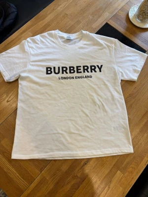 Vit Burberry t-shirt   - Säljer en klassisk vit t-shirt från Burberry med stort svart logotryck på bröstet där det står 'BURBERRY LONDON ENGLAND'. T-shirten har rund hals och korta ärmar. Tillverkad i mjuk bomull för en skön känsla.