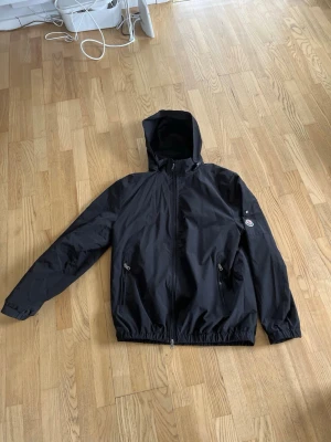 Svart vindjacka från Moncler XL - Snygg svart vindjacka från Moncler med huva och dragkedja framtill. Två fickor med dragkedja och Moncler-logga på ärmen. Tillverkad i lätt, vindtätt material som passar perfekt för blåsiga dagar. Lägger man telefonen mot loggan kommer man in på moncler appen.