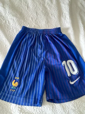 Blå fotbollsshorts Frankrike Nike - Säljer ett par blå fotbollsshorts från Nike med Frankrikes landslagsmärke och nummer 10 på ena benet. Shortsen har vita och röda tunna ränder, elastisk midja och är tillverkade i ett lätt och ventilerande material. Perfekta för träning eller match!