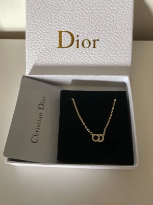 Guldhalsband med pärlor från Dior - Snyggt halsband från Christian Dior i guldtonad metall med ett gnistrande hängsmycke format som Diors ikoniska logga, dekorerat med små pärlor. Kedjan har en rund bricka med Dior-gravyr vid låset. Perfekt accessoar för att lyfta din outfit.