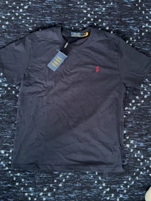 Mörkblå t-shirt från Polo Ralph Lauren - Snygg mörkblå t-shirt från Polo Ralph Lauren med klassisk röd logga broderad på bröstet. Tillverkad i mjuk bomull och har en normal passform med korta ärmar och rund hals. Perfekt till jeans eller shorts för en clean look.