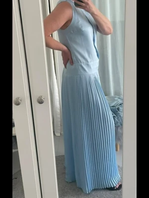 Ljusblå långklänning - Säljer en elegant ljusblå långklänning med plisserad kjol och knappar framtill. Klänningen är ärmlös och har en markerad midja som ger en snygg siluett. Perfekt för sommarens alla tillfällen!