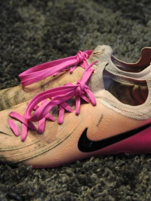 Nike Phantom GX 2 - Säljer ett par Nike fotbollsskor i ljus orange med svarta swoosh-loggor och starkt rosa skosnören. Skorna har en slimmad form och är tillverkade i ett lätt material som passar perfekt för match eller träning. Storlek 44. Missfärgningen är pga användning på konstgräs under några få veckor bara. 
