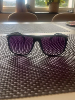  Solglasögon  - Snygga solglasögon med svart båge och rektangulär form. Glasen är lila-tonade vilket ger en cool kontrast till den svarta ramen. Perfekt accessoar för dig som vill sticka ut lite extra i solen.