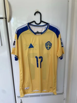 Svensk landslagströja Adidas gul/blå - Gul och blå fotbollströja från Adidas med Sveriges landslagsemblem på bröstet och nummer 17 i blått framtill. Tröjan har korta ärmar, blå detaljer på axlar och ärmslut samt tryckta sponsormärken på ärmarna. Perfekt för fotbollsfans!