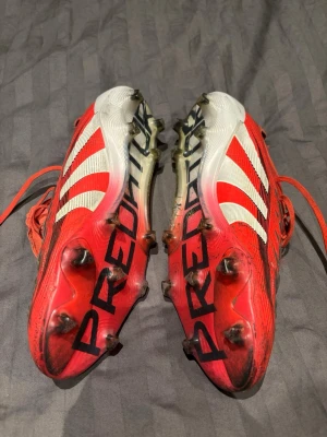 Adidas Predator röda fotbollsskor stl 46,2/3 - Säljer ett par röda Adidas Predator fotbollsskor med vita och svarta detaljer. Skorna har klassiska tre ränder på sidan, snörning och dobbar för bra grepp på planen. Tillverkade i syntetmaterial och designade för maximal bollkontroll och komfort. OBS ena gummibandet till tungan är borta.
