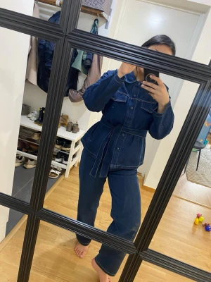 Mörkblå jeans set med bälte 50% bomull - Säljer en snygg mörkblå jeansjumpsuit med markerad midja och matchande bälte. Jumpsuiten har långa ärmar, stora bröstfickor med knappar och klassisk krage. Materialet är kraftigt denim med kontrastsömmar för en cool look.