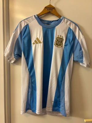 Argentina fotbollströja Adidas - Säljer en klassisk Argentina landslagströja från Adidas med blå och vita ränder, guldfärgat AFA-märke och Adidas-logga på bröstet. Tillverkad i lätt och ventilerande material, perfekt för fotboll eller träning.
