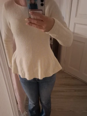 Krämvit peplumstickad tröja - Säljer en krämvit stickad tröja med peplumformad midja och långa ärmar. Tröjan har rund halsringning och är tillverkad i ett mjukt, ribbat material som ger en snygg siluett. Perfekt för dig som gillar stilrena och feminina plagg.