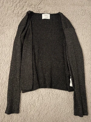 Glittrig svart kofta från Zara - Snygg svart kofta från Zara med glittriga inslag och ribbad struktur. Koftan har långa ärmar och knäppning framtill med två diskreta knappar. Perfekt för att addera lite extra shine till din outfit. Passar strl S