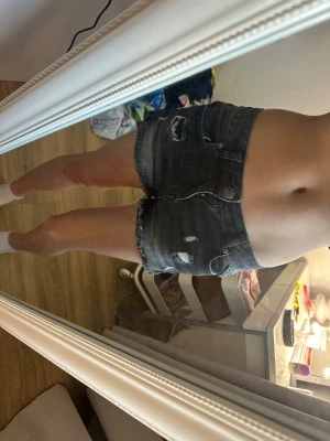 Mörkgrå jeansshorts med slitningar  - Säljer ett par mörkgrå jeansshorts med råa kanter och slitna detaljer. Klassisk femficksmodell med knapp och dragkedja framtill. Snygg tvättad look och kort passform, perfekta till sommaren.